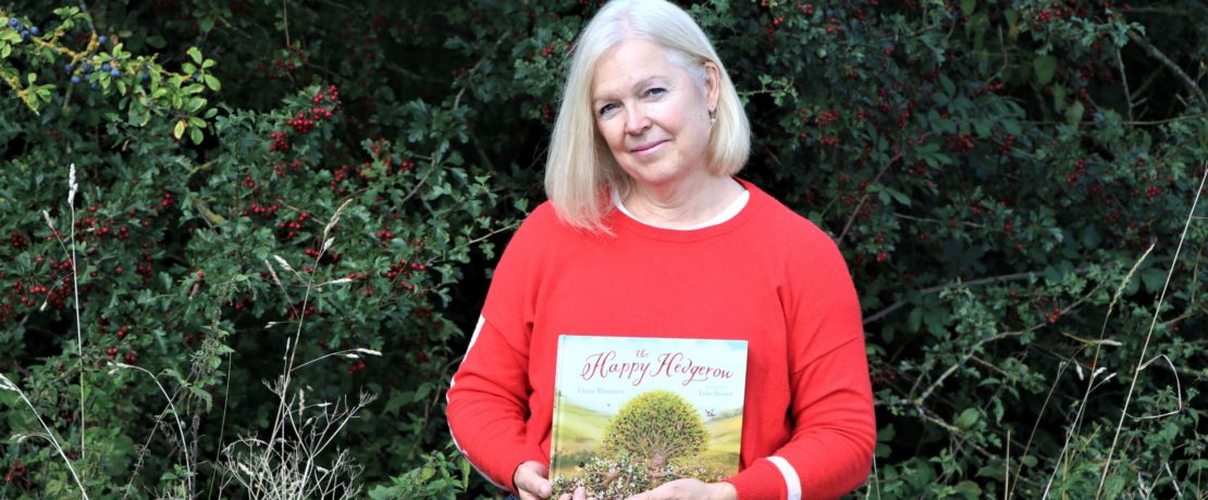 the author of The Happy Hedgerow holding her book, standing in front of a lush green hedge