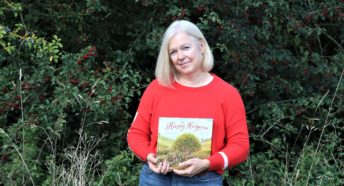 the author of The Happy Hedgerow holding her book, standing in front of a lush green hedge