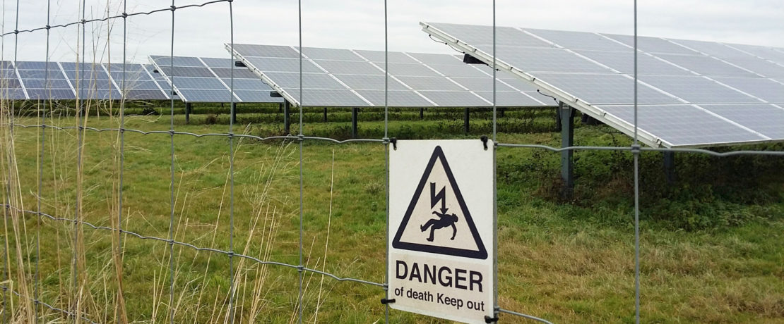 rows of glass photovoltaic panels behind a metal fence with a warning sign that says danger of death keep out