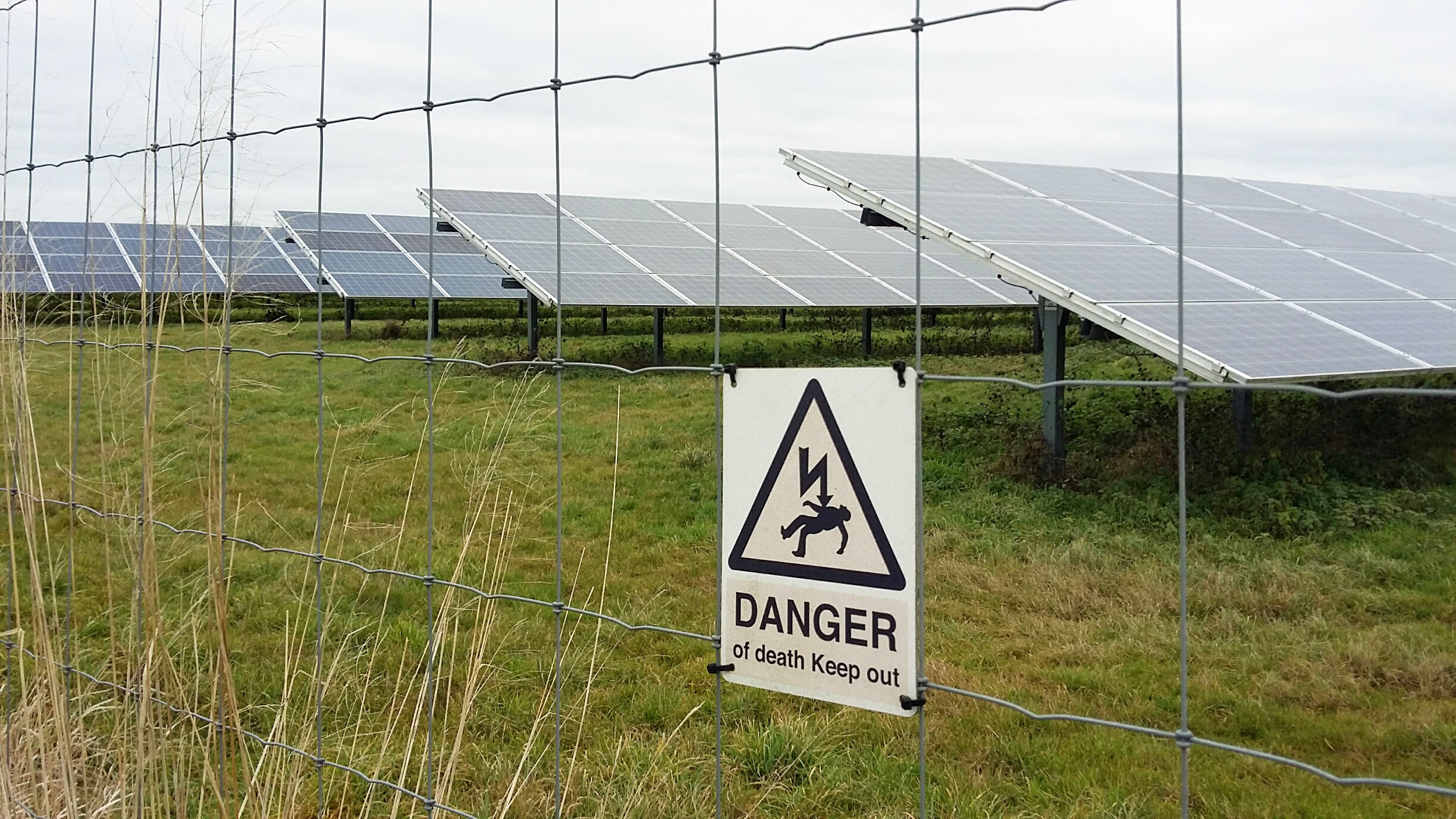 rows of glass photovoltaic panels behind a metal fence with a warning sign that says danger of death keep out