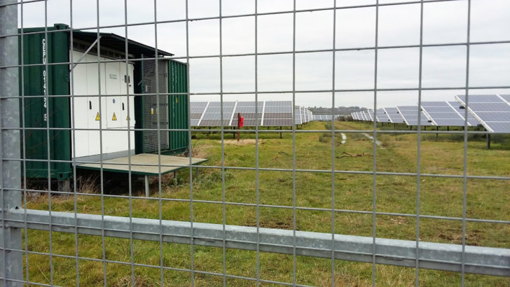 many rows of solar photovolteic panels and a transformer in large metal container behind a metal fence