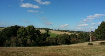 long range view over green grassy fields, woodlands and distant hills with a blue sky and puffy white clouds overhead