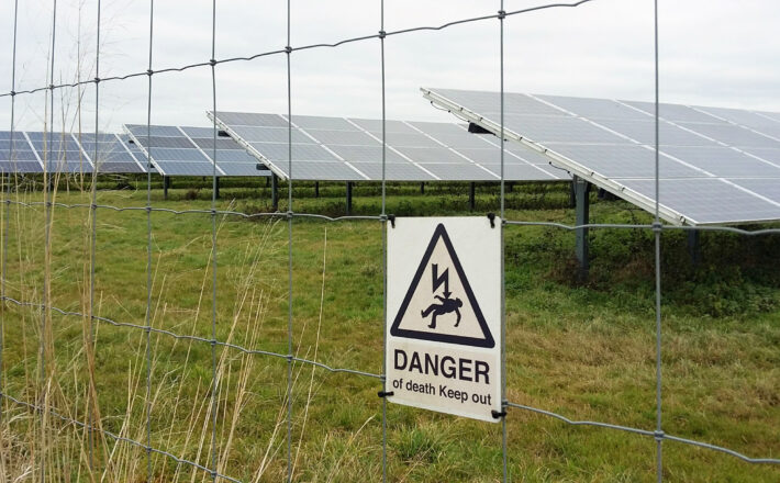rows of glass photovoltaic panels behind a metal fence with a warning sign that says danger of death keep out